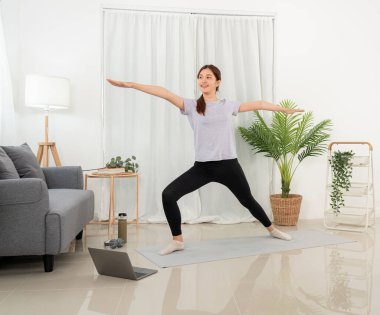 Young woman is taking yoga lesson online on laptop and standing to practicing yoga with pose warrior two exercise while workout on mat in her living room at home.