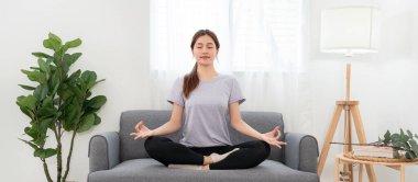 Young woman is relaxing into the lotus position for meditation on couch before workout yoga exercise in her living room at home.
