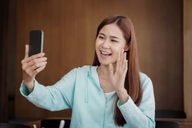 Female student using smartphone to greeting with classmate on video call while studying online learning to preparing for examination in library.