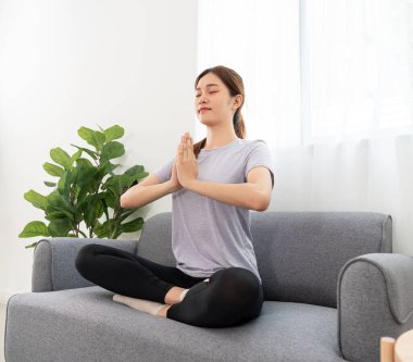 Young woman is relaxing into the lotus position with namaste gesture for meditation on couch before workout yoga exercise in her living room at home.
