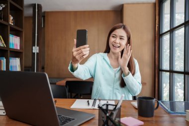Female student using smartphone to greeting with classmate on video call while studying online learning to preparing for examination in library.
