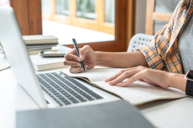Young woman student in headphone is learning lessons online on laptop with lecturer and taking notes on notebook to preparing for examination in college.