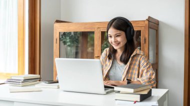 Young woman student in headphone is learning lessons online on laptop with lecturer and taking notes on notebook to preparing for examination in college.