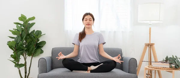 Young woman is relaxing into the lotus position for meditation on couch before workout yoga exercise in her living room at home.