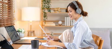 Kadın serbest yazar, müzik dinlemek için kulaklık takarken ve merkez ofisteki oturma odasındaki masada çalışırken dizüstü bilgisayardaki iş verilerini okuyor ve not defterine bilgi yazıyor..