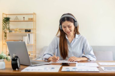 Kadın serbest yazar, müzik dinlemek için kulaklık takarken dizüstü bilgisayarda iş verilerini okuyor ve not defterine bilgi yazıyor ve merkez ofisteki masada çalışıyor..