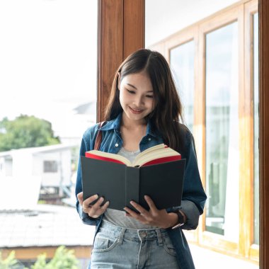 Young woman student is standing near the window and reading literature to relaxation while taking a break between lesson classes in college.