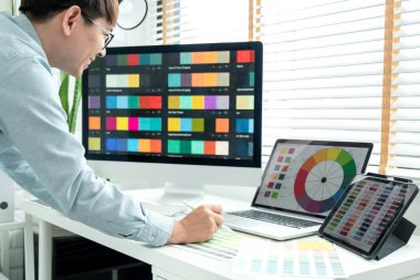 Male graphic designer is standing to choosing color swatch samples on multiple screens and working graphic design with technology while sketching on tablet digital in modern office.
