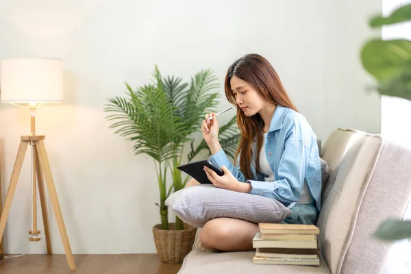 Young woman is analyzing strategy goal of business and taking notes on tablet while working on comfortable the couch in living room at home.