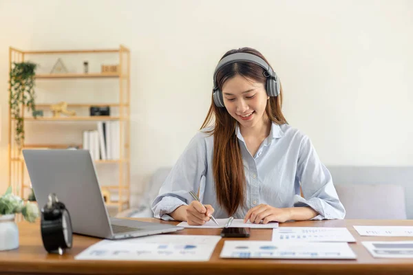 Female freelance is reading business data on laptop and writing information on notebook while wearing headphone to listening music and working on the table in living room at home office.