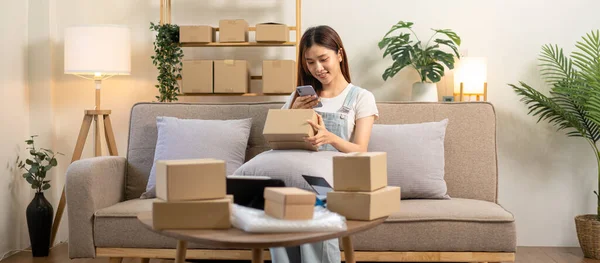 Young entrepreneur is holding cardboard boxes to taking a picture to sending customer while sitting on comfortable couch and working in living room at home office.
