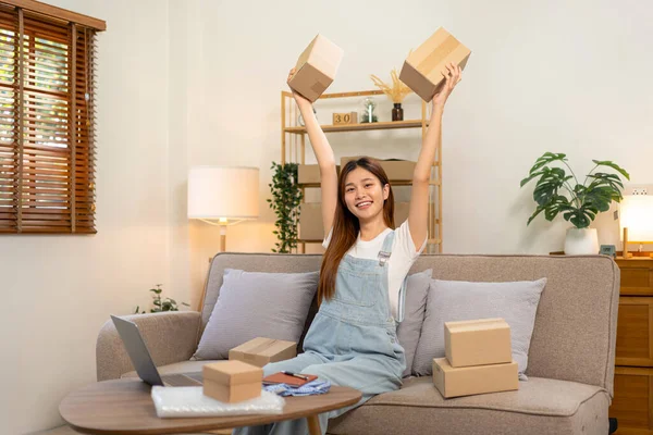 Young entrepreneur is holding cardboard boxes and raising arms after successfully in sale products while sitting on comfortable the couch and working in living room at home office.
