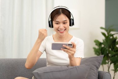 Genç kız evde uzaktan öğrenim gördükten sonra oturma odasındaki kanepede oturup rahatlamak için kulaklık takıyor ve akıllı telefondan oyun oynuyor..