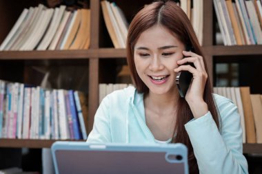 Kız öğrenci kütüphanede tabletteki verileri okurken akıllı telefondan sınıf arkadaşlarıyla ödev ödevlerini konuşuyor ve anlatıyor.