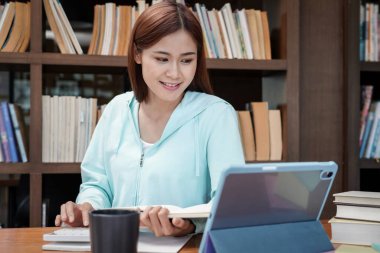 Kadın öğrenci, kütüphanede ders kitabı okuyup veri ararken finans ve muhasebe ödevi yapmak için hesap makinesi kullanıyor.
