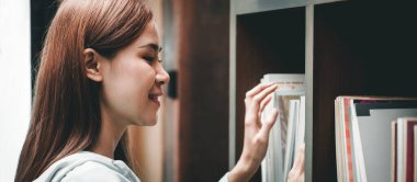 Female student is standing in front of the bookshelf to choosing textbook or literature in library between take a break from studying lessons class at university.