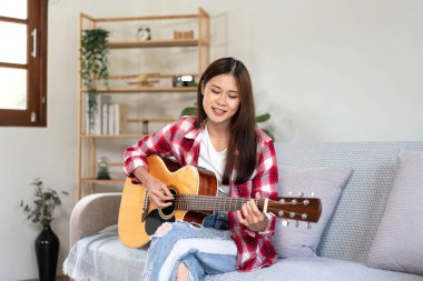 Genç kadın evdeki kanepede rahat bir şekilde otururken gitar çalıyor ve şarkı söylemeye çalışıyor..