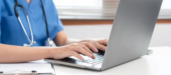 Mavi üniformalı veteriner kadın veteriner kliniğinde çalışırken evcil hayvan bilgilerini aramak ve tedavi verilerini daktilo etmek için laptop kullanıyor..