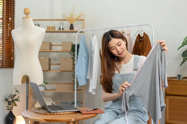 Female online seller is using laptop to broadcast live on Vlog for introduce fashions while measuring the chest size of clothes to showing for customer and selling online clothing on social media.