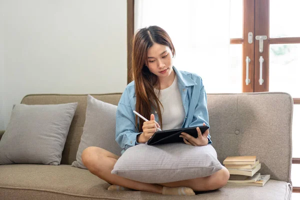 年轻女性在家里客厅舒适的沙发上工作时 正在分析公司的战略目标 并在平板电脑上记笔记 — 图库照片