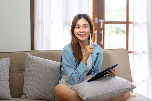 Young woman is holding pen and tablet to thinking about strategy goal of business while working on comfortable the couch in living room at home.