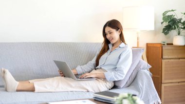 Serbest kadın, iş verilerini okuyup dizüstü bilgisayarda bilgi yazarken aynı zamanda kanepede rahat bir şekilde oturuyor ve merkez ofisteki oturma odasında çalışıyor..