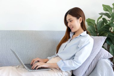 Serbest kadın, iş verilerini okuyup dizüstü bilgisayarda bilgi yazarken aynı zamanda kanepede rahat bir şekilde oturuyor ve merkez ofisteki oturma odasında çalışıyor..