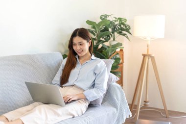 Serbest kadın, iş verilerini okuyup dizüstü bilgisayarda bilgi yazarken aynı zamanda kanepede rahat bir şekilde oturuyor ve merkez ofisteki oturma odasında çalışıyor..