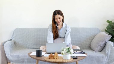 Serbest çalışan kadın, iş verilerini okuyup dizüstü bilgisayarında bilgi yazarken aynı zamanda yastıkla rahat bir şekilde kanepede oturuyor ve merkez ofisteki oturma odasında çalışıyor..
