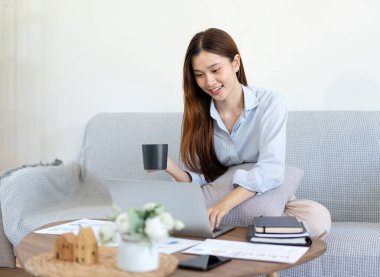 Kadın serbest yazar dizüstü bilgisayarda bilgi yazıyor ve kanepede yastıkla rahat otururken kahve içiyor ve merkez ofisteki oturma odasında çalışıyor..