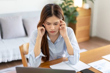 Kadın serbest çalışan, dizüstü bilgisayarda iş raporlarını okurken ve merkez ofisteki oturma odasındaki masada çalışırken strateji planını düşünmekten dolayı stresli ve baş ağrısı çekiyor..