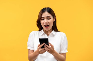 Beyaz kısa kollu gömlek giyen genç Asyalı kadın sarı arka planda izole edilmiş akıllı telefona bakarken şok ve heyecan hissediyordu..