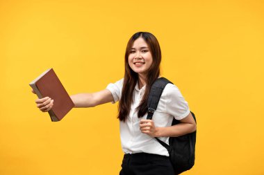 Sırtında kitap ve omuz çantası tutan kız öğrenci gülümsüyor ve üniversiteye hazırlanıyor sarı arka planda izole bir şekilde..