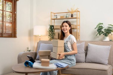 Genç girişimci elinde karton kutularla müşterilere mektup ve ev teslimatı hazırlarken aynı zamanda kanepede oturuyor ve merkez büroda oturma odasında çalışıyor..