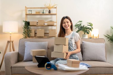 Genç girişimci elinde karton kutularla müşterilere mektup ve ev teslimatı hazırlarken aynı zamanda kanepede oturuyor ve merkez büroda oturma odasında çalışıyor..