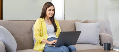 Genç bir kadın rahat bir koltukta oturmuş dizüstü bilgisayarını bacaklarına takıyor ve oturma odasında çalışırken iş raporu yazıyor..