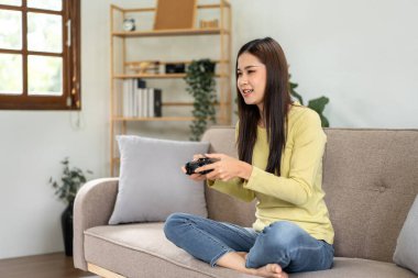 Genç Asyalı kadın evdeki oturma odasındaki büyük rahat kanepede otururken oyun kumandası joystick 'ini oyun oynamak için kullanıyor..