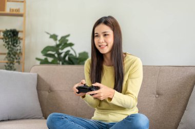 Genç Asyalı kadın evdeki oturma odasındaki büyük rahat kanepede otururken oyun kumandası joystick 'ini oyun oynamak için kullanıyor..
