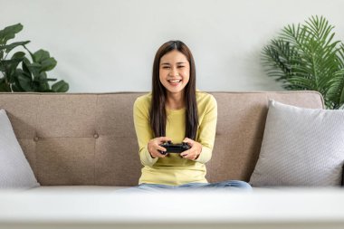 Genç Asyalı kadın evdeki oturma odasındaki büyük rahat kanepede otururken oyun kumandası joystick 'ini oyun oynamak için kullanıyor..