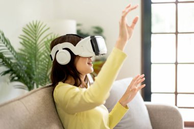 Sanal gerçeklik gözlüğü takan Asyalı genç bir kadın sanal gerçeklik gözlüğü takıyor ve parmaklarıyla havaya dokunmak ve oturma odasındaki kanepede oturmak için el ele tutuşurken sanal deneyimle oyunlar oynuyor..