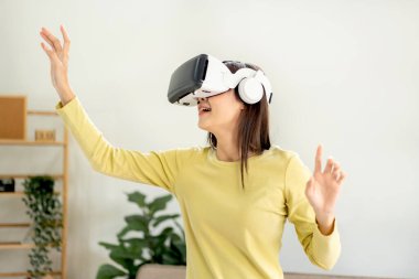 Sanal gerçeklik gözlüğü takan Asyalı genç bir kadın sanal gerçeklik gözlüğü takıyor ve parmaklarıyla havaya dokunmak ve evdeki oturma odasında durmak için el ele tutuşarak sanal deneyimle oyunlar oynuyor..