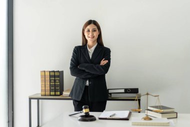 İş kadını avukat kadın masada pirinç pullar ve adalet çekiciyle çalışırken kolları çapraz duruyor ve hukuk bürosundaki kameraya bakıp gülümsüyor..