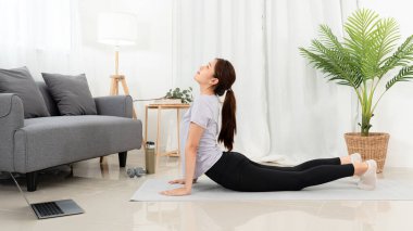 Genç bir kadın laptopta yoga dersi izliyor. Evindeki oturma odasında minder üzerinde kobra pozu vererek yoga yapıyor..