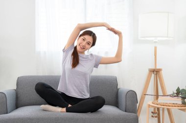 Genç kadın, evindeki oturma odasında yoga yaptıktan sonra koltukta rahatlamak için kollarını esnetiyor..