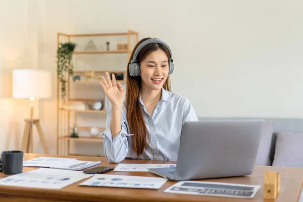 Female freelance is using laptop and wearing headphone to meeting video conference with colleague while working on the table in living room at home office.