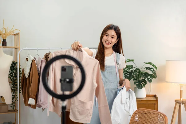 Female online seller is using smartphone to broadcast live on Vlog for introduce fashions while showing shirt to presenting new collection and selling online clothing on social media.