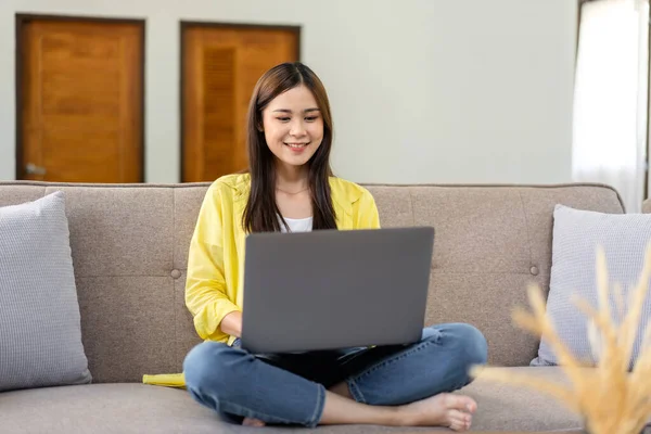 Young woman is sitting with squat on comfortable couch and putting laptop on the legs to typing business report while working in living room at home.