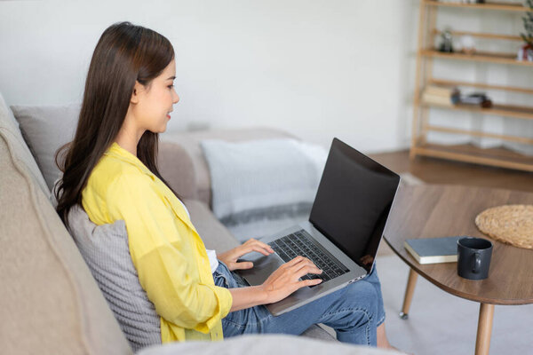 Молодая женщина сидит со скрещенными ногами на удобном диване и кладет ноутбук на ноги, чтобы печатать бизнес-отчет, работая в гостиной на дому.