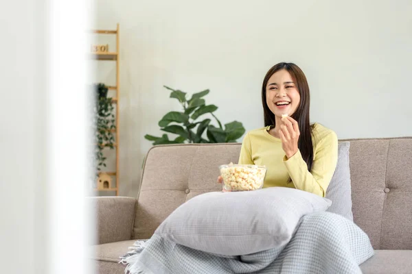 Genç Asyalı kadın yastığa bir kase patlamış mısır koyup patlamış mısır yerken büyük rahat koltukta oturup evdeki oturma odasında televizyon seyrediyor..
