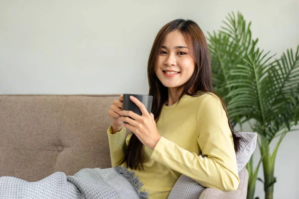 年轻的亚洲女人正在喝咖啡 躺在舒适的大沙发上 在家客厅里看电视上的电影 — 图库照片